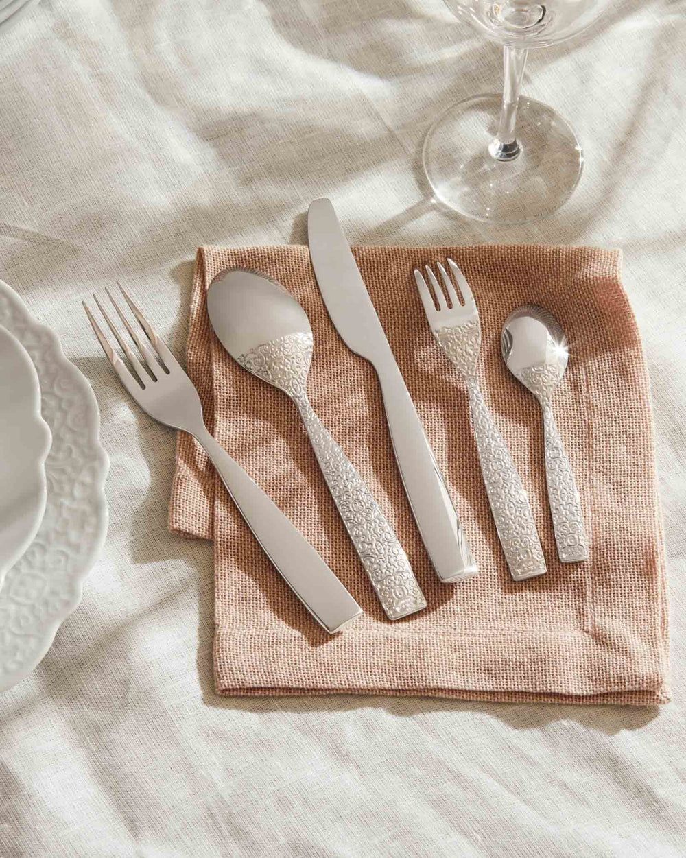 Five-piece silver cutlery set by Alessi featuring ornate embossed handles on a pink napkin.