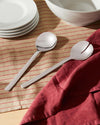 Two shiny Ovale silver salad servers by Alessi, one slotted, resting on a striped linen tablecloth with white plates.