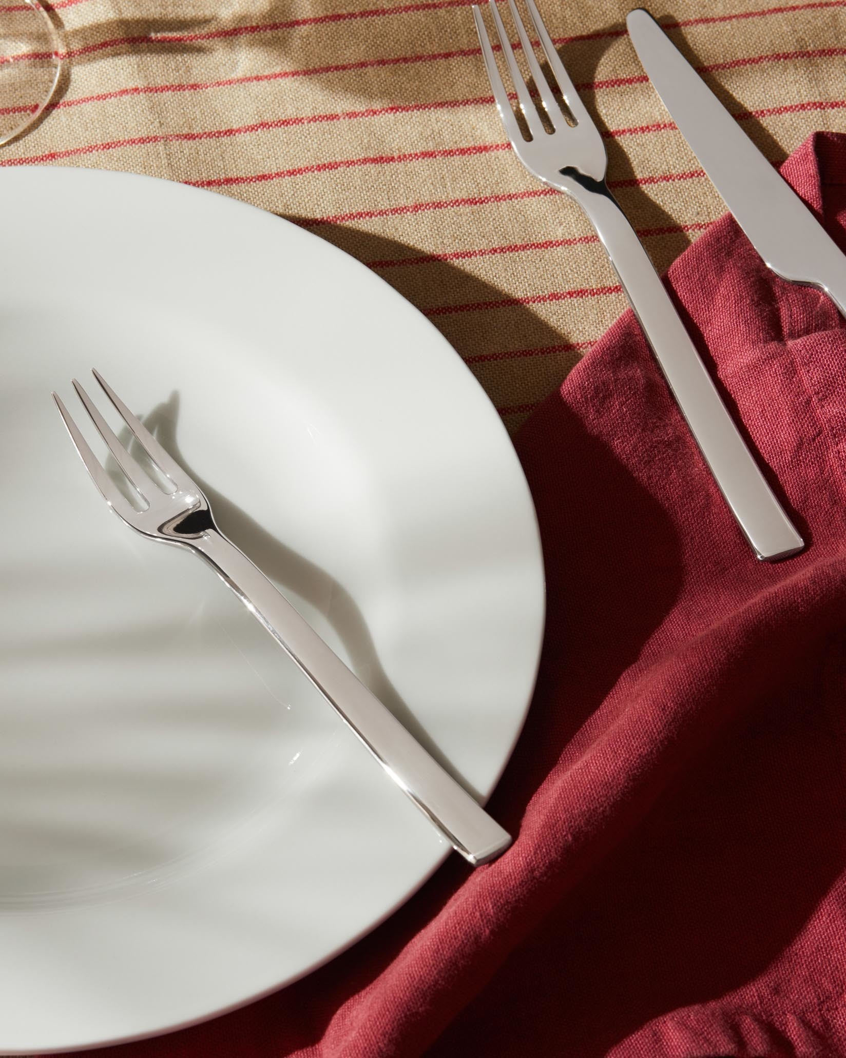 Silver Ovale cutlery set by Alessi, with a fork and knife on a white plate, red napkin, and striped tablecloth.