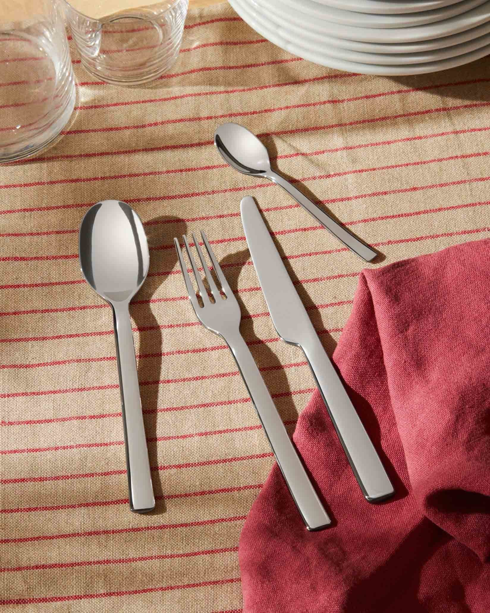 Silver-toned Ovale cutlery set by Alessi, including fork, knife, and spoons, on a striped linen tablecloth with a red napkin.