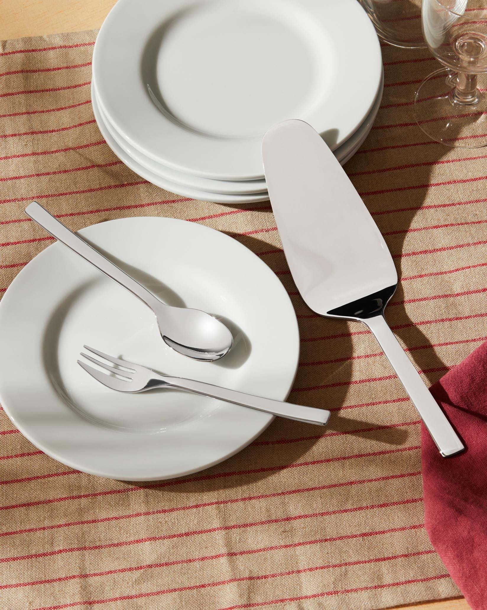Sleek silver cutlery set by Alessi with fork, spoon, and cake server on a white plate, next to a red napkin and striped tablecloth.