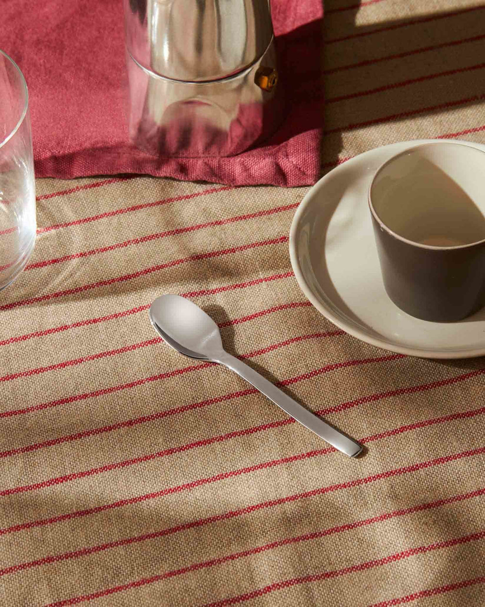 Silver Ovale spoon by Alessi on a striped tablecloth, next to a coffee cup and glass.