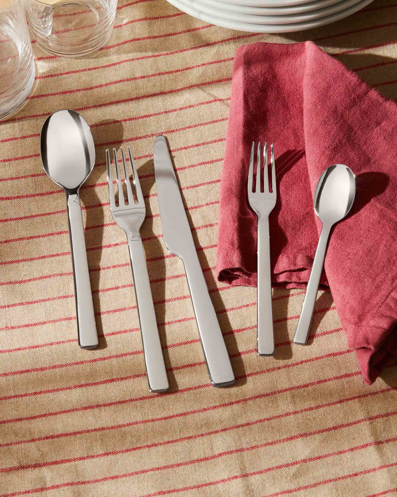 Stainless steel Ovale cutlery set by Alessi, elegantly laid on a beige striped tablecloth with a red napkin and white plates.
