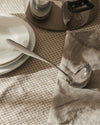 Silver stainless steel ladle by Alessi, resting on a beige checkered tablecloth beside white plates and a grey linen napkin.