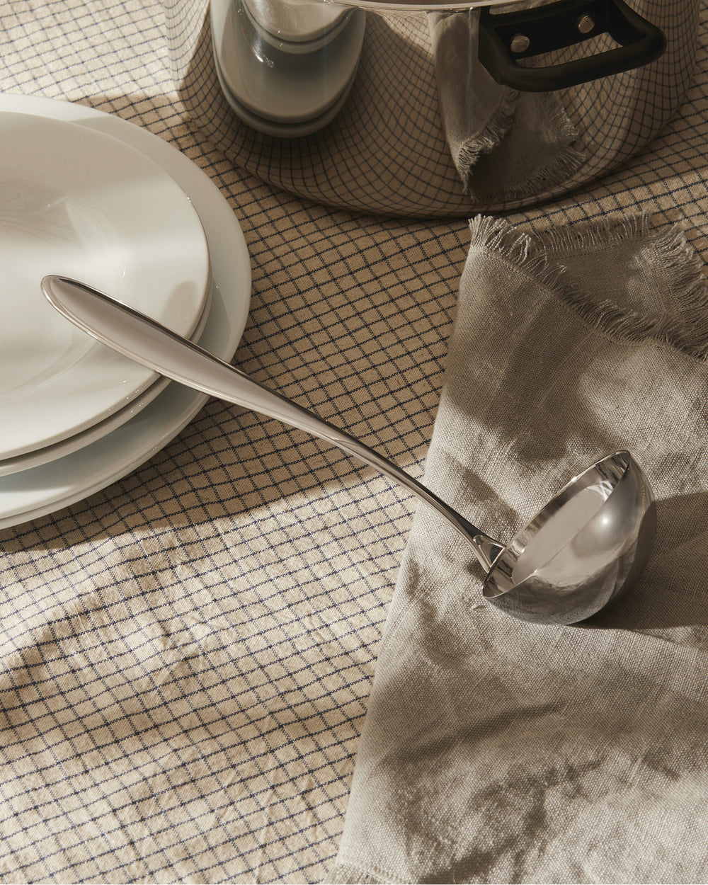 Silver stainless steel ladle by Alessi, resting on a beige checkered tablecloth beside white plates and a grey linen napkin.