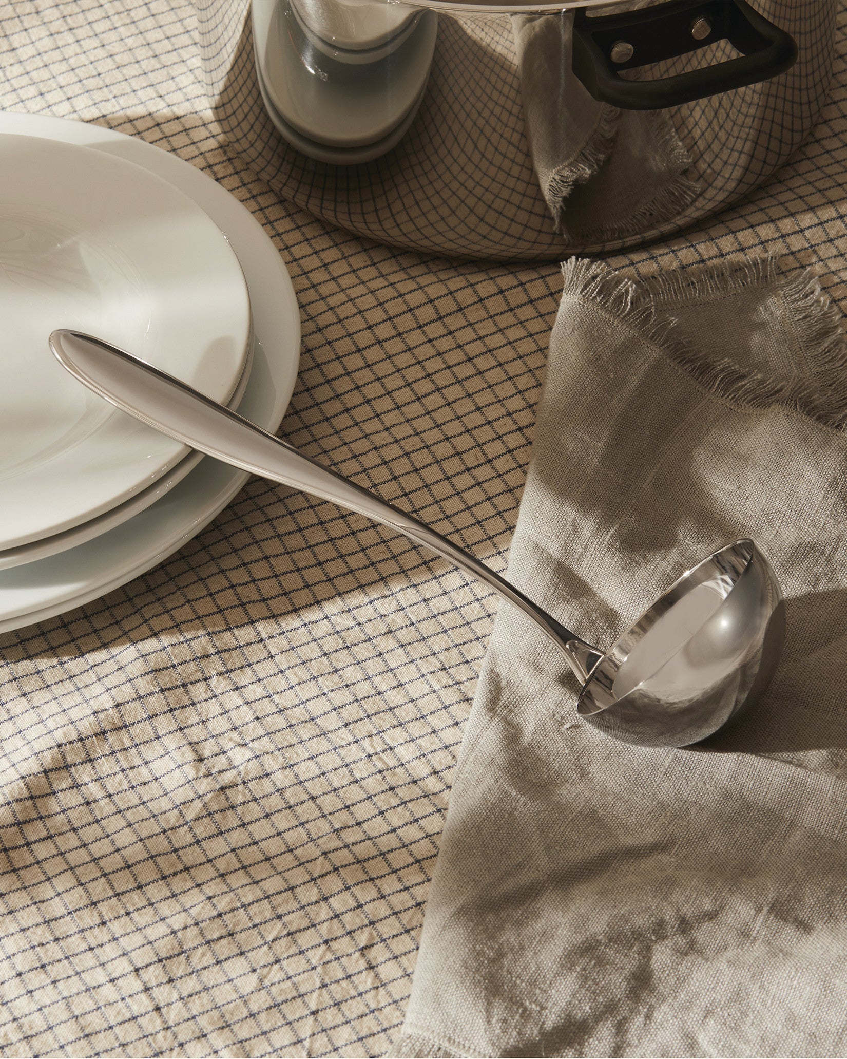 Silver stainless steel ladle by Alessi, resting on a beige checkered tablecloth beside white plates and a grey linen napkin.