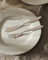 Sleek silver fork and knife by Alessi, resting on a white plate on a checkered tablecloth with a brown linen napkin.