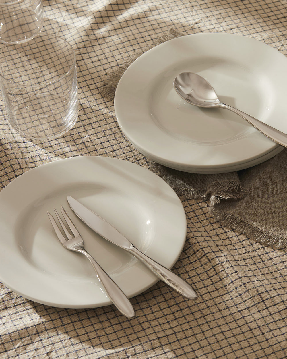 White ceramic tableware set by Alessi, with silver cutlery and clear glasses on a beige checkered tablecloth.