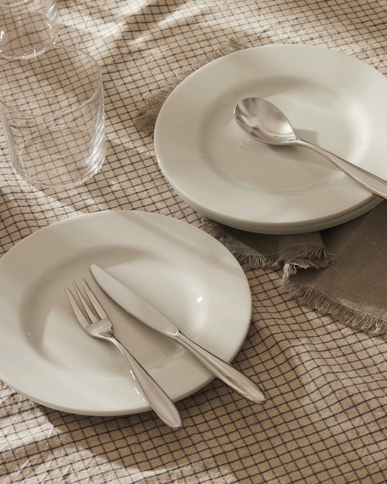 White ceramic tableware set by Alessi, with silver cutlery and clear glasses on a beige checkered tablecloth.