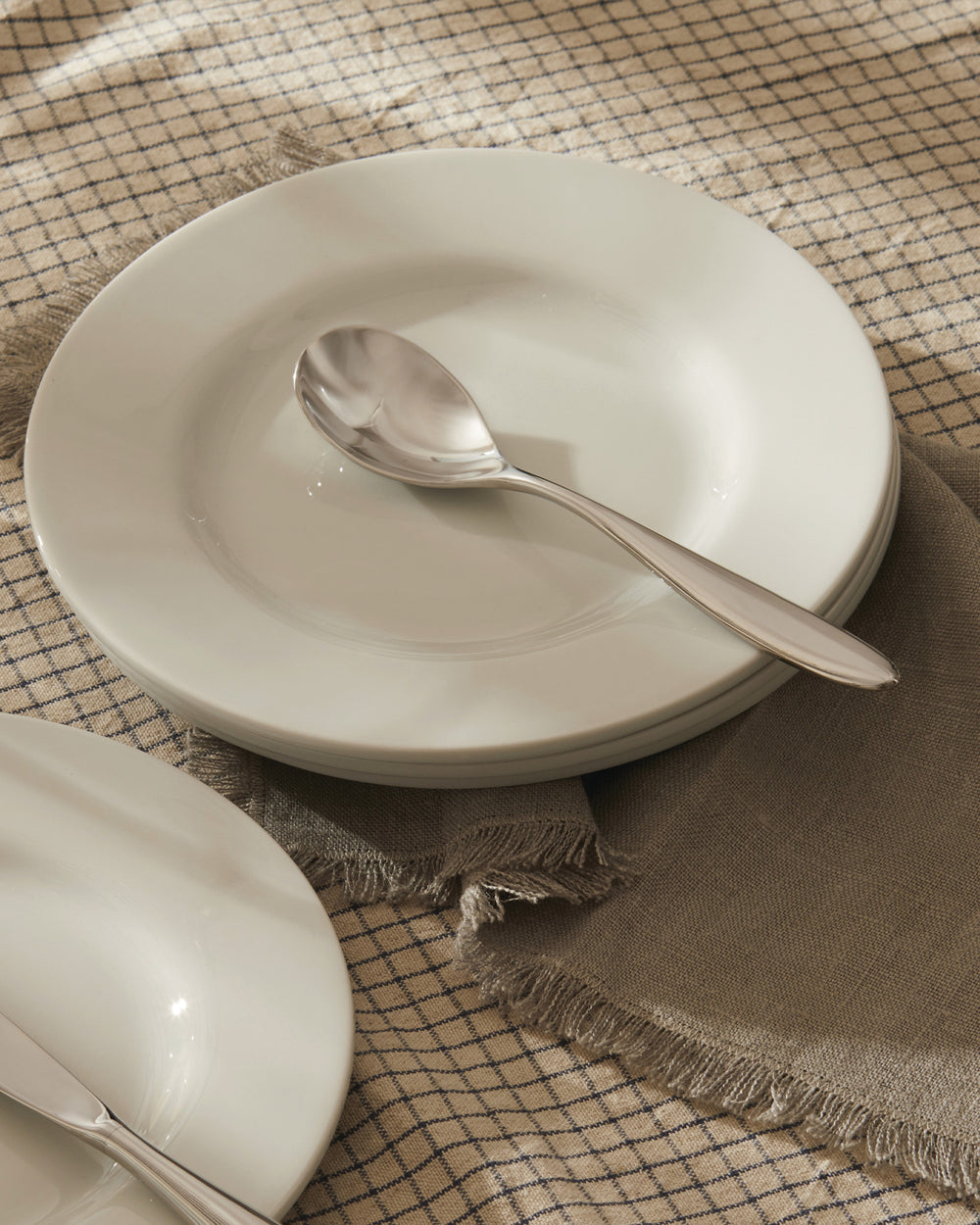 White ceramic dinner plates by Alessi, stacked with a spoon on a checkered tablecloth and grey napkin.