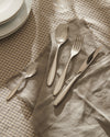 Polished silver cutlery set by Alessi, four pieces on a frayed grey linen napkin and plaid tablecloth.