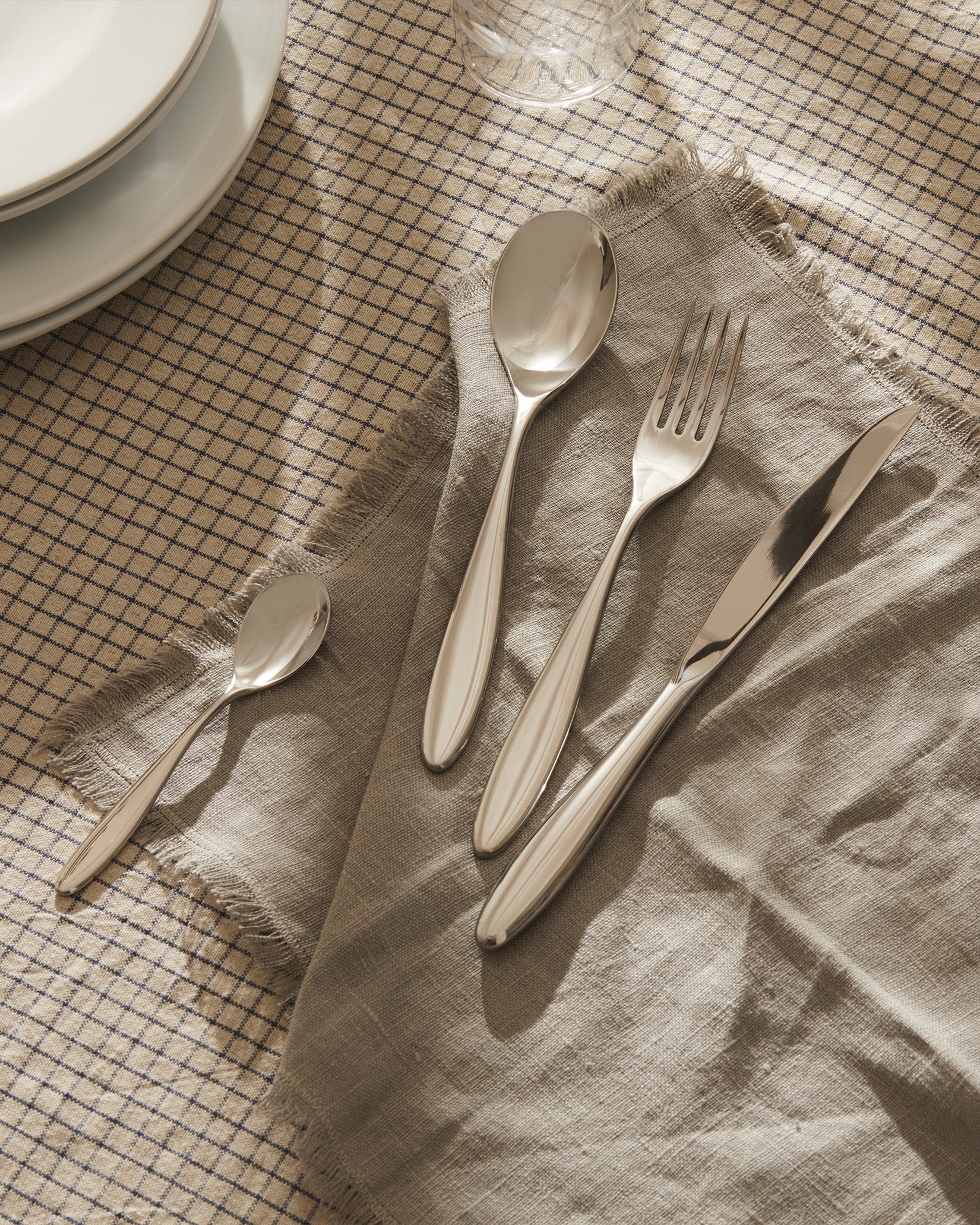 Polished silver cutlery set by Alessi, four pieces on a frayed grey linen napkin and plaid tablecloth.