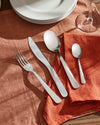 Silver stainless steel cutlery set by Alessi, with a sleek, faceted design on an orange linen placemat.