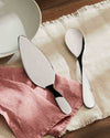 Polished silver serving utensils by Alessi, including a cake server and large spoon, on pink and beige linen cloths.