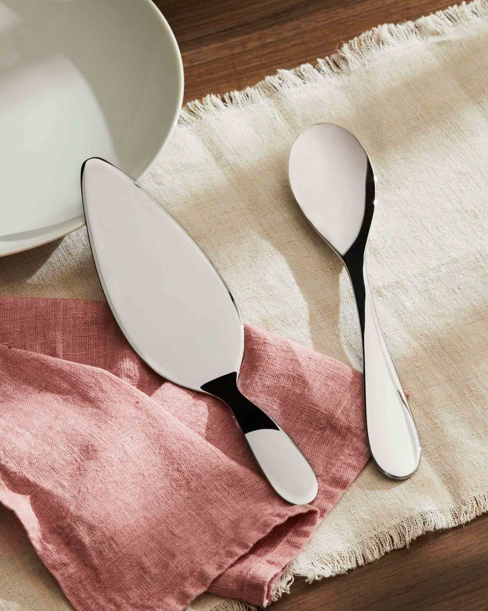 Polished silver serving utensils by Alessi, including a cake server and large spoon, on pink and beige linen cloths.