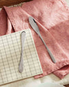 Sleek silver-toned eat.it spreader by Alessi on a pink linen napkin next to a white grid-patterned plate.