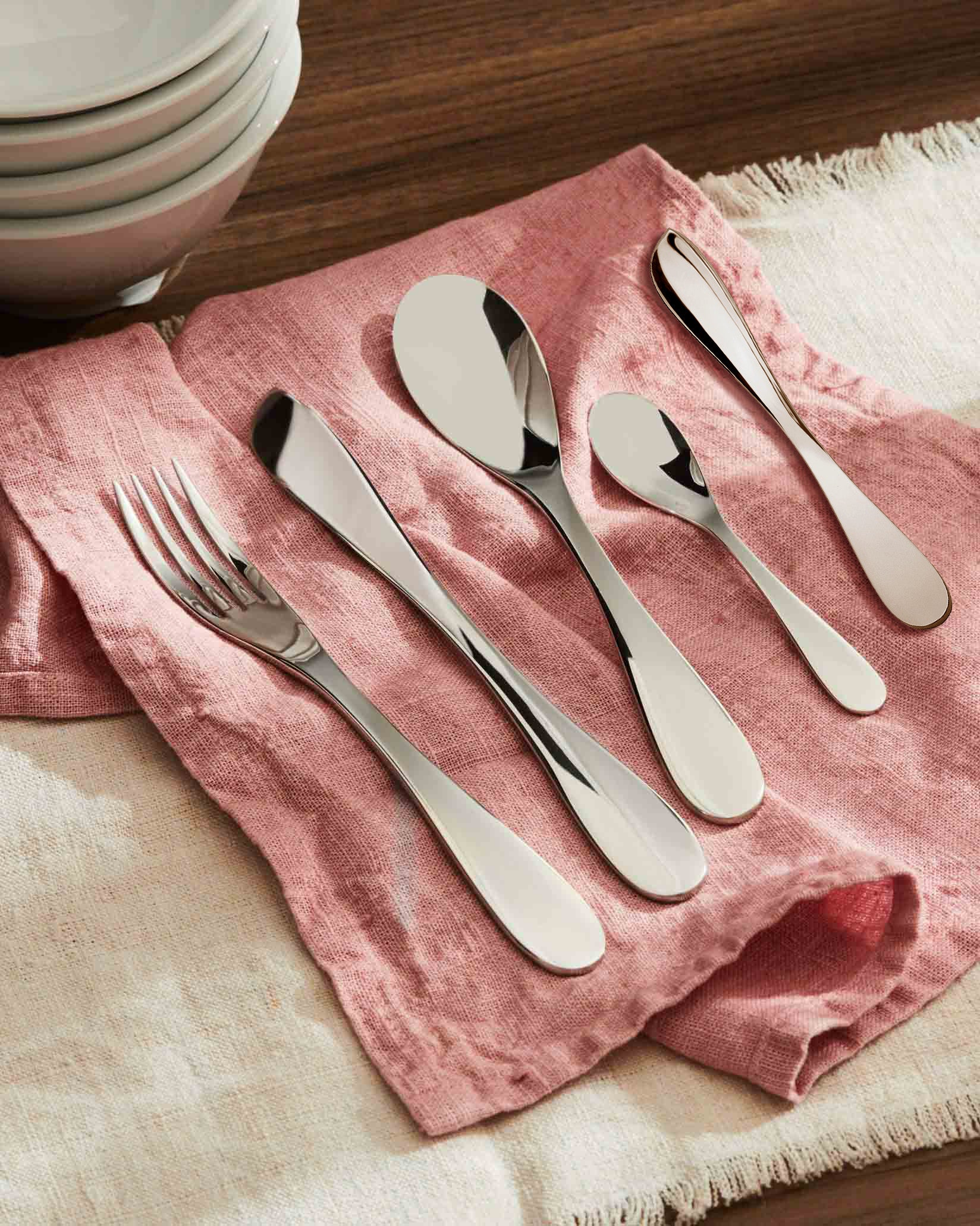 Silver stainless steel cutlery set by Alessi, elegantly laid on a pink linen napkin next to stacked white bowls.
