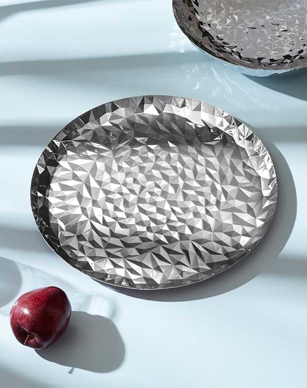 Silver metallic platter by Alessi with a faceted geometric pattern, next to a red apple.