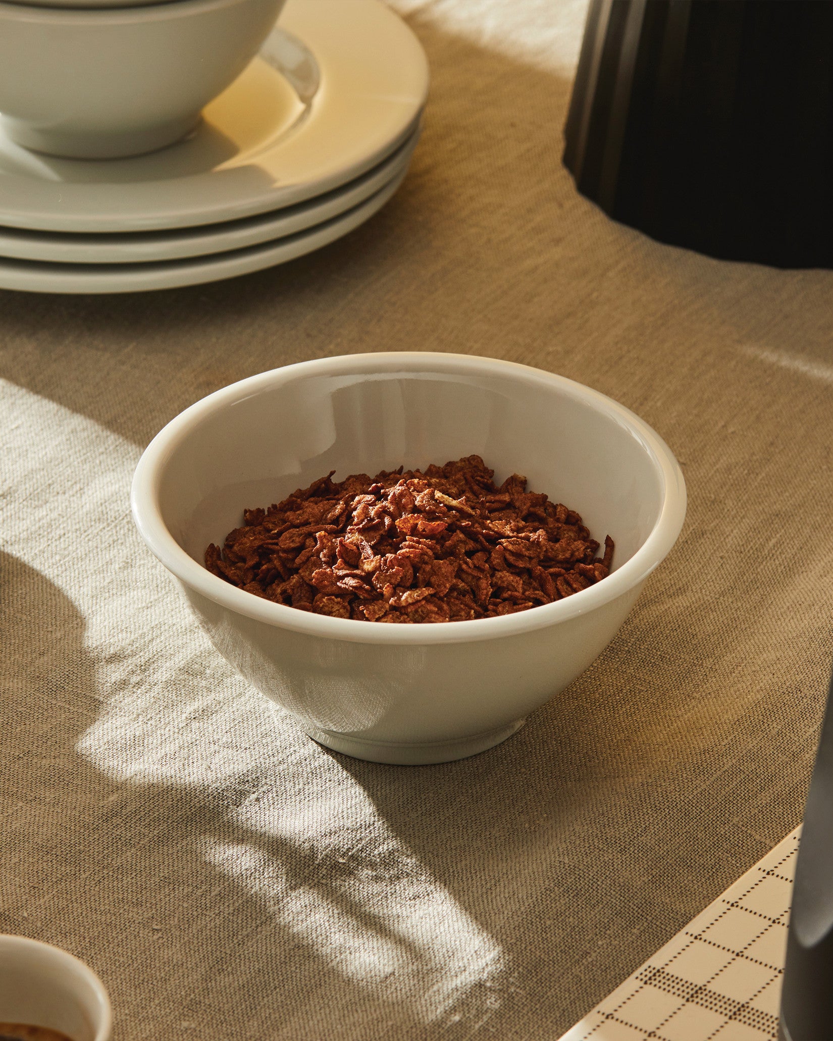 White ceramic bowl by Alessi filled with chocolate cereal, on a beige linen tablecloth next to stacked plates.