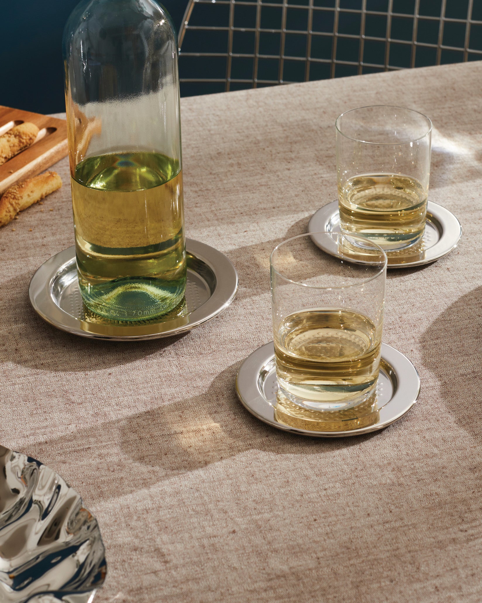 Silver metallic round coasters by Alessi with a dimpled pattern, displayed under a bottle and glasses on a textured tablecloth.