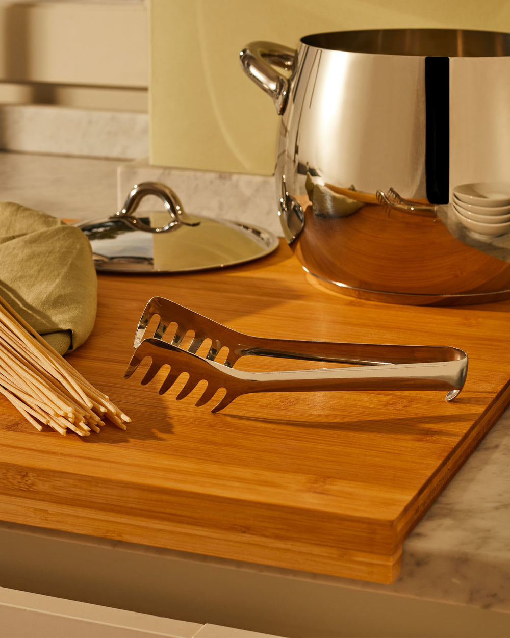 Stainless steel pasta tongs by Alessi, open and resting on a wooden cutting board with uncooked spaghetti.