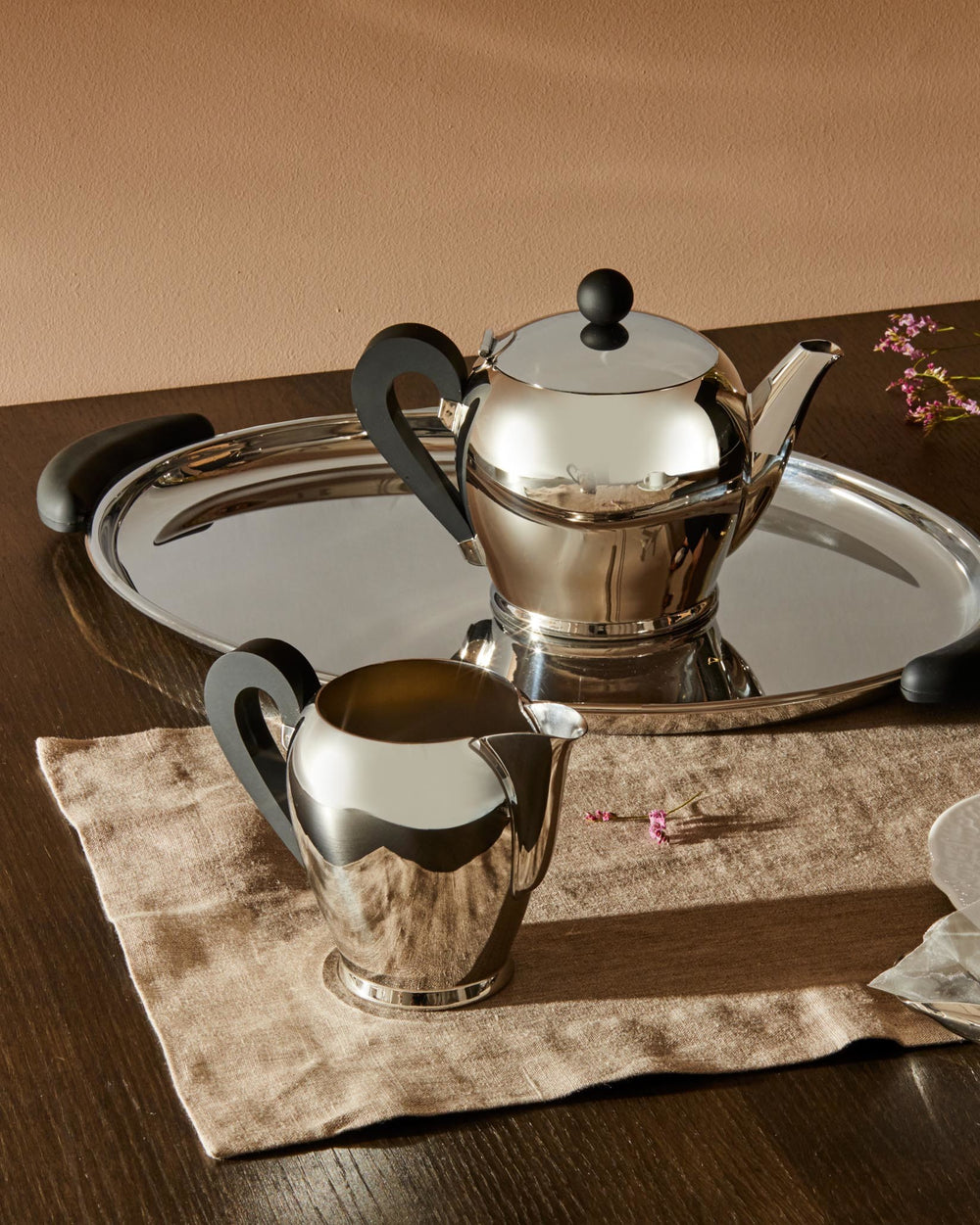 Shiny silver Bombé teapot by Alessi with black handles, a matching creamer, and a silver tray on a brown linen cloth.