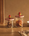 Glass storage jars by Alessi with pink lids, yellow discs, and red spherical knobs, filled with cinnamon and dried oranges on a wooden surface.