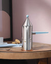 Silver conical coffee maker with a light blue handle by Alessi, featuring a copper base, placed on a wooden table.
