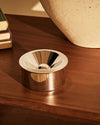 Silver metallic ashtray by Alessi, with a polished, reflective finish and a concave top, on a wooden surface.
