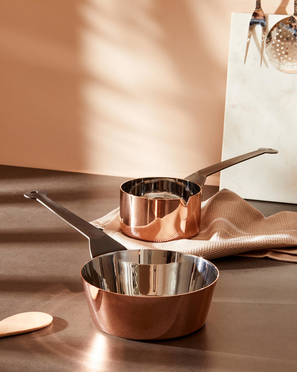 Two copper and stainless steel casseroles by Alessi with long dark handles, on a kitchen counter.