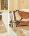 Silver stainless steel Moka pot by Alessi being assembled by hands on a marble countertop.