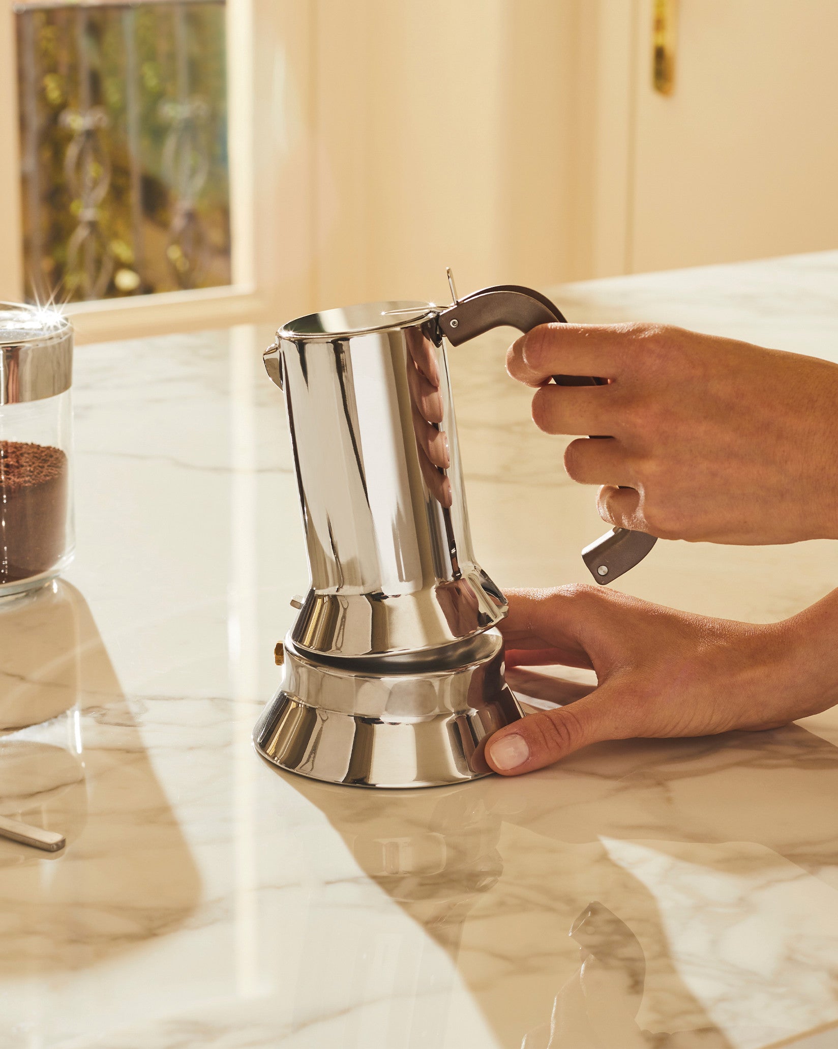 Silver stainless steel Moka pot by Alessi being assembled by hands on a marble countertop.