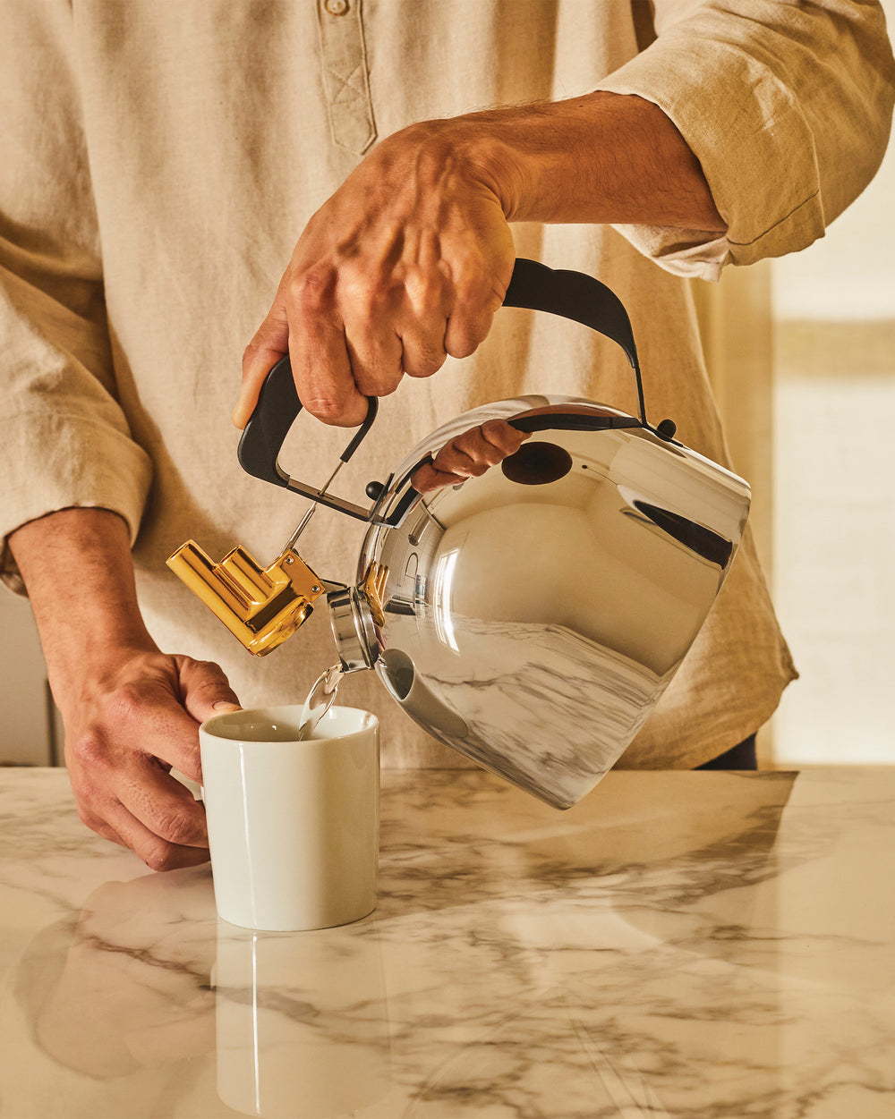 Chrome kettle by Alessi with a gold whistle and black handle, pouring water into a white mug on a marble counter.