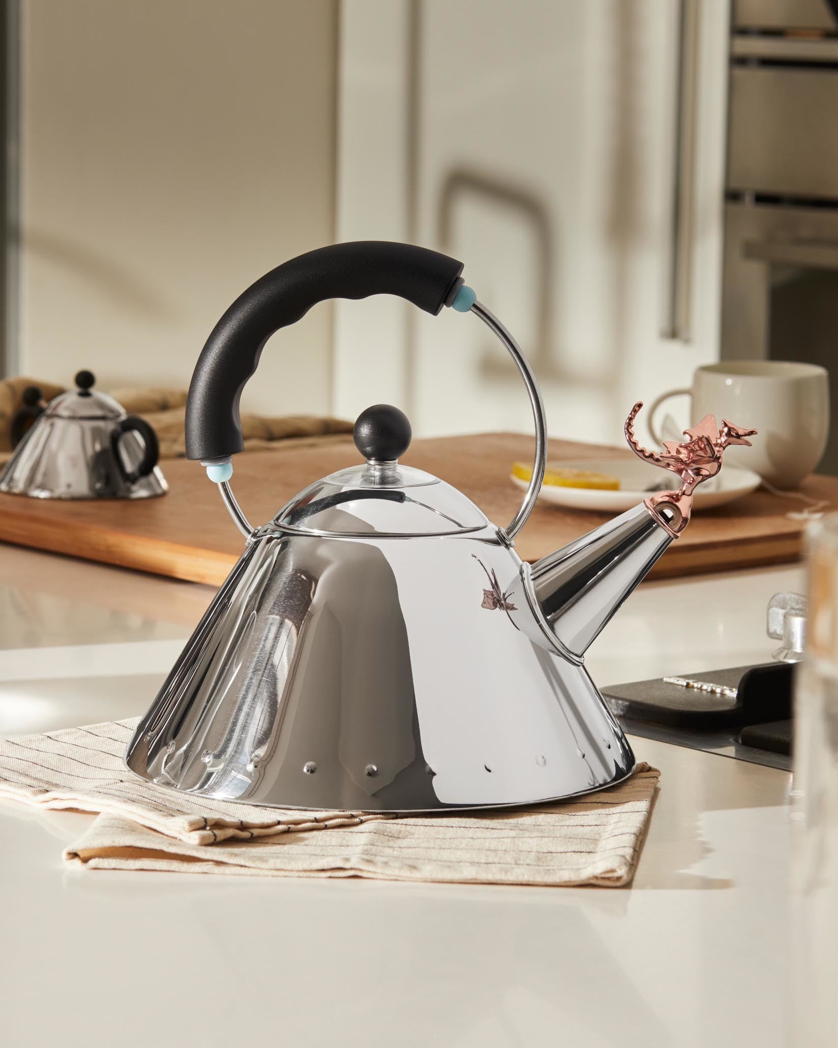 Shiny stainless steel whistling tea kettle by Alessi, featuring a rose gold dragon-shaped whistle and a black handle.