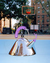 Reflective chrome conical kettle by Alessi with a purple handle and person-shaped whistle, on a blue court.