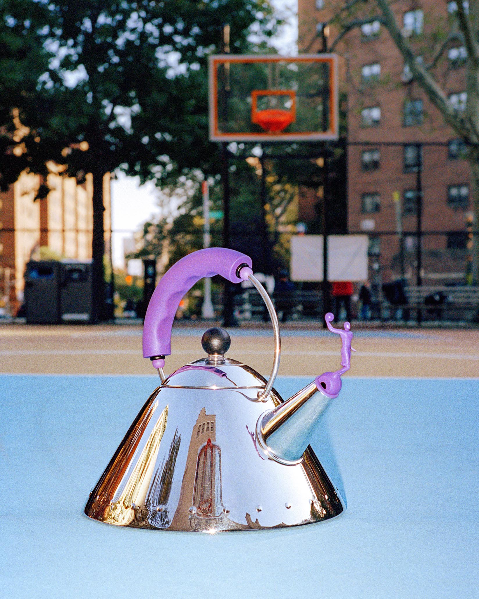 Reflective chrome conical kettle by Alessi with a purple handle and person-shaped whistle, on a blue court.