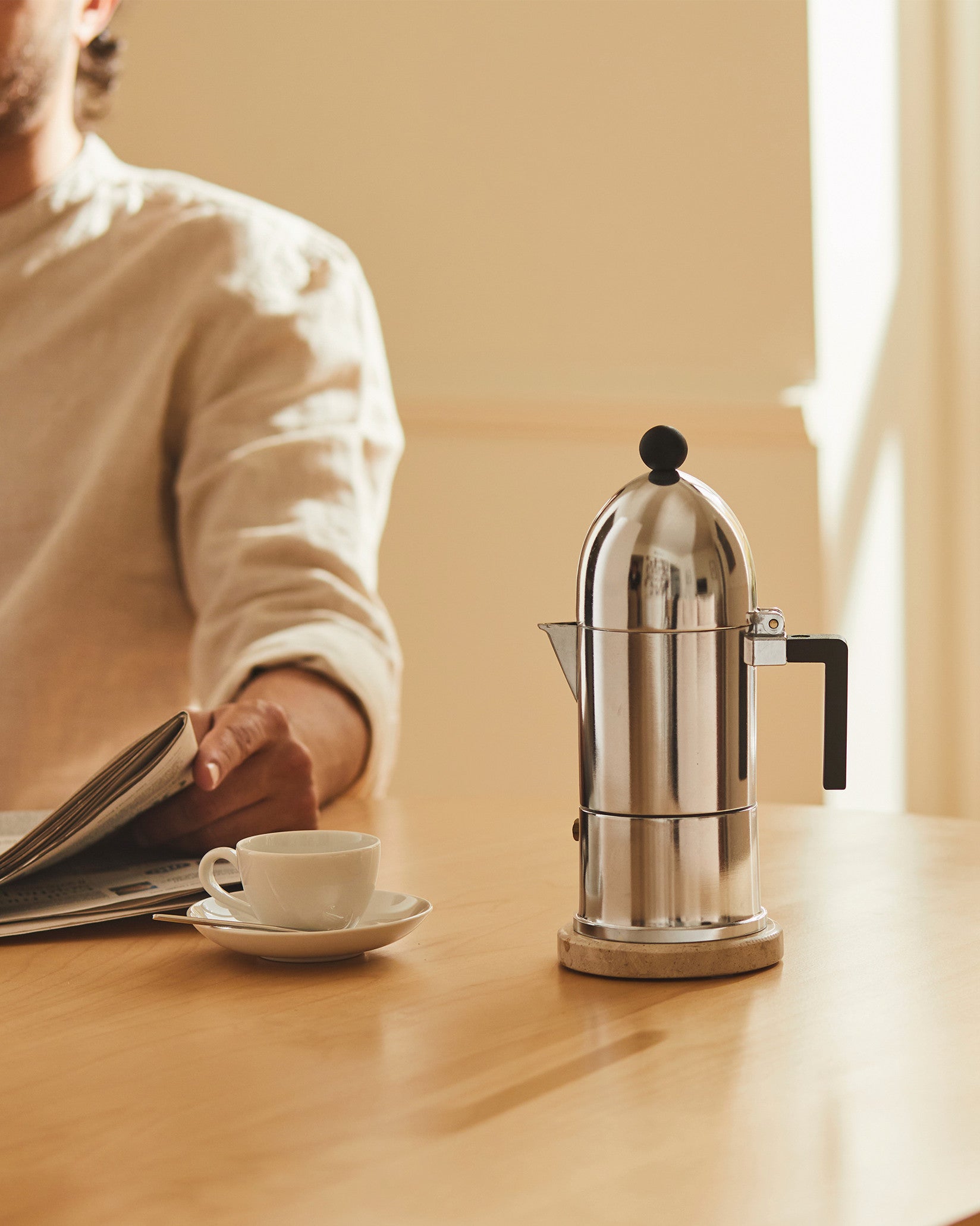 Silver polished moka pot by Alessi with a black handle and knob, next to a white coffee cup and a person reading a newspaper.