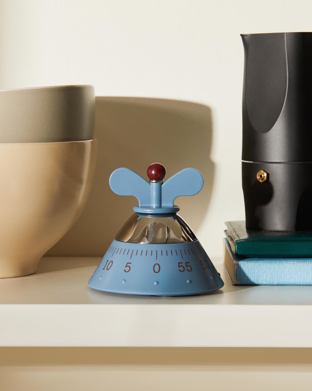 Light blue and chrome Kitchen Timer by Alessi with a red knob and wing-shaped handles, on a shelf with ceramic bowls and a moka pot.