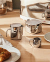Silver-toned stainless steel creamer with a hinged lid by Alessi, displayed alongside a matching square sugar bowl and a small creamer cup on a wooden table.