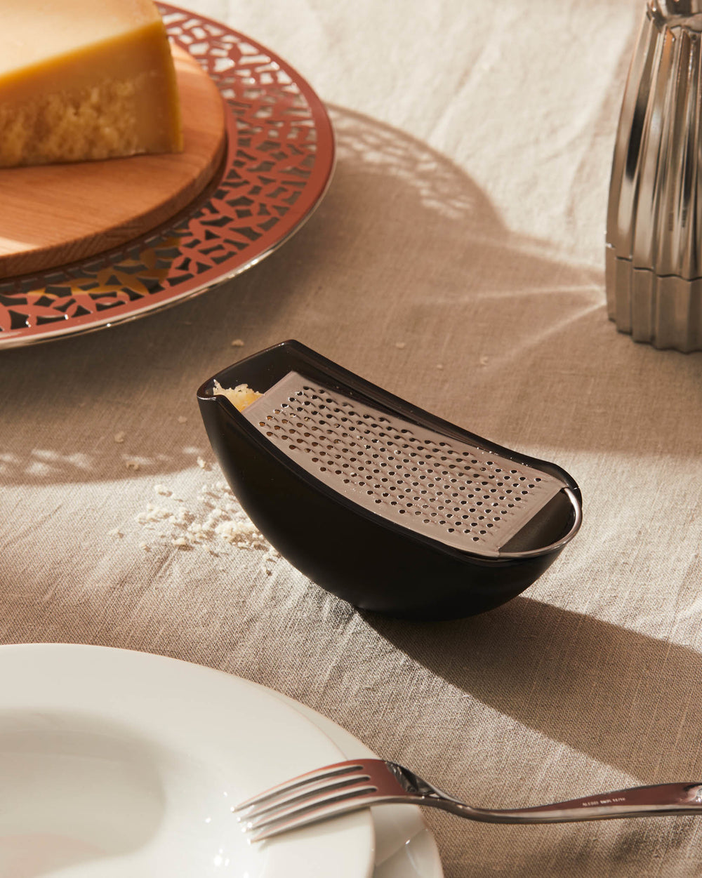 Black tabletop cheese grater by Alessi, with a stainless steel grating surface and grated cheese on a linen tablecloth.