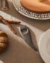 Dark gray devil-themed bottle opener by Alessi, resting on a linen tablecloth with breadsticks and a cheese board.