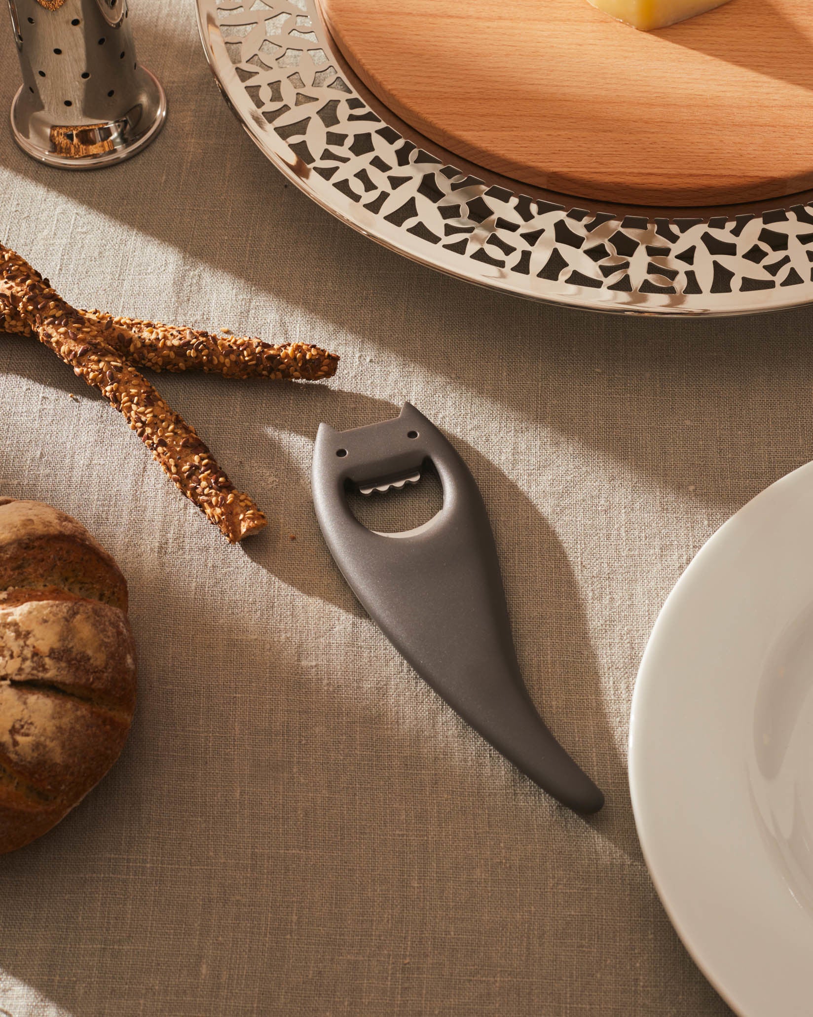 Dark gray devil-themed bottle opener by Alessi, resting on a linen tablecloth with breadsticks and a cheese board.