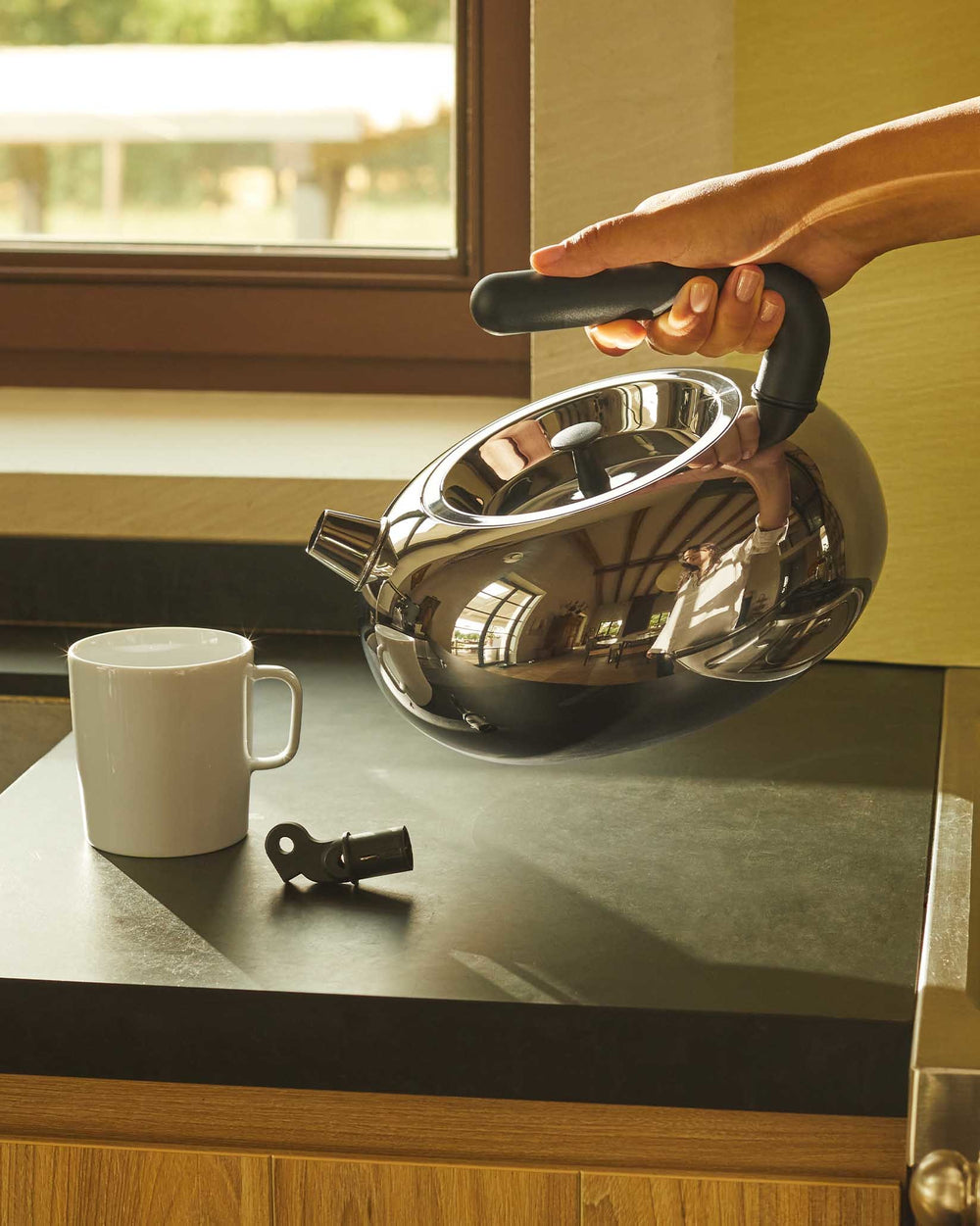Silver stainless steel Bulbul kettle by Alessi, held by a hand over a white mug on a dark countertop.