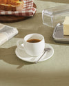 White ceramic espresso cup and saucer set by Alessi, with a spoon, on a light green tablecloth.