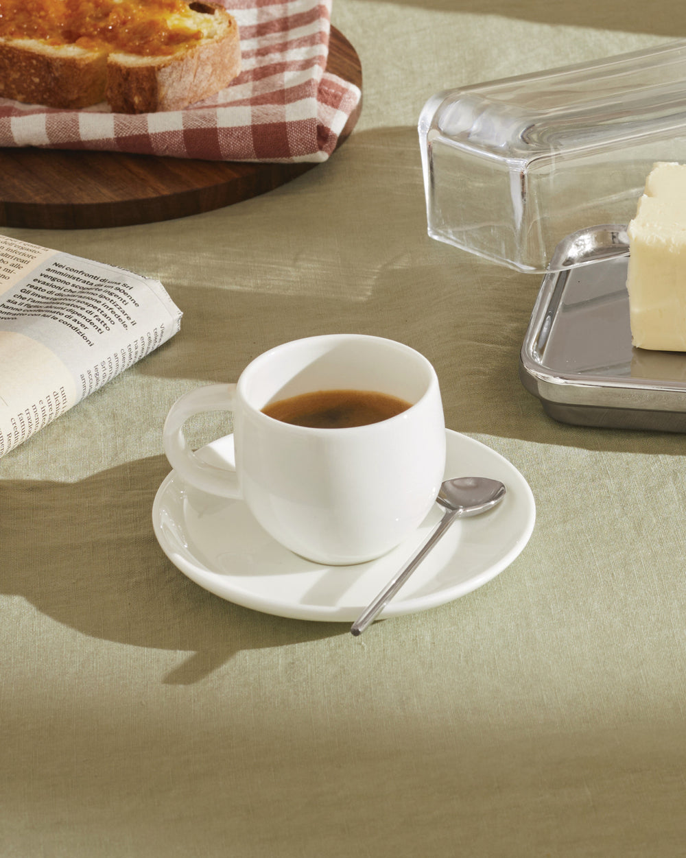 White ceramic espresso cup and saucer set by Alessi, with a spoon, on a light green tablecloth.