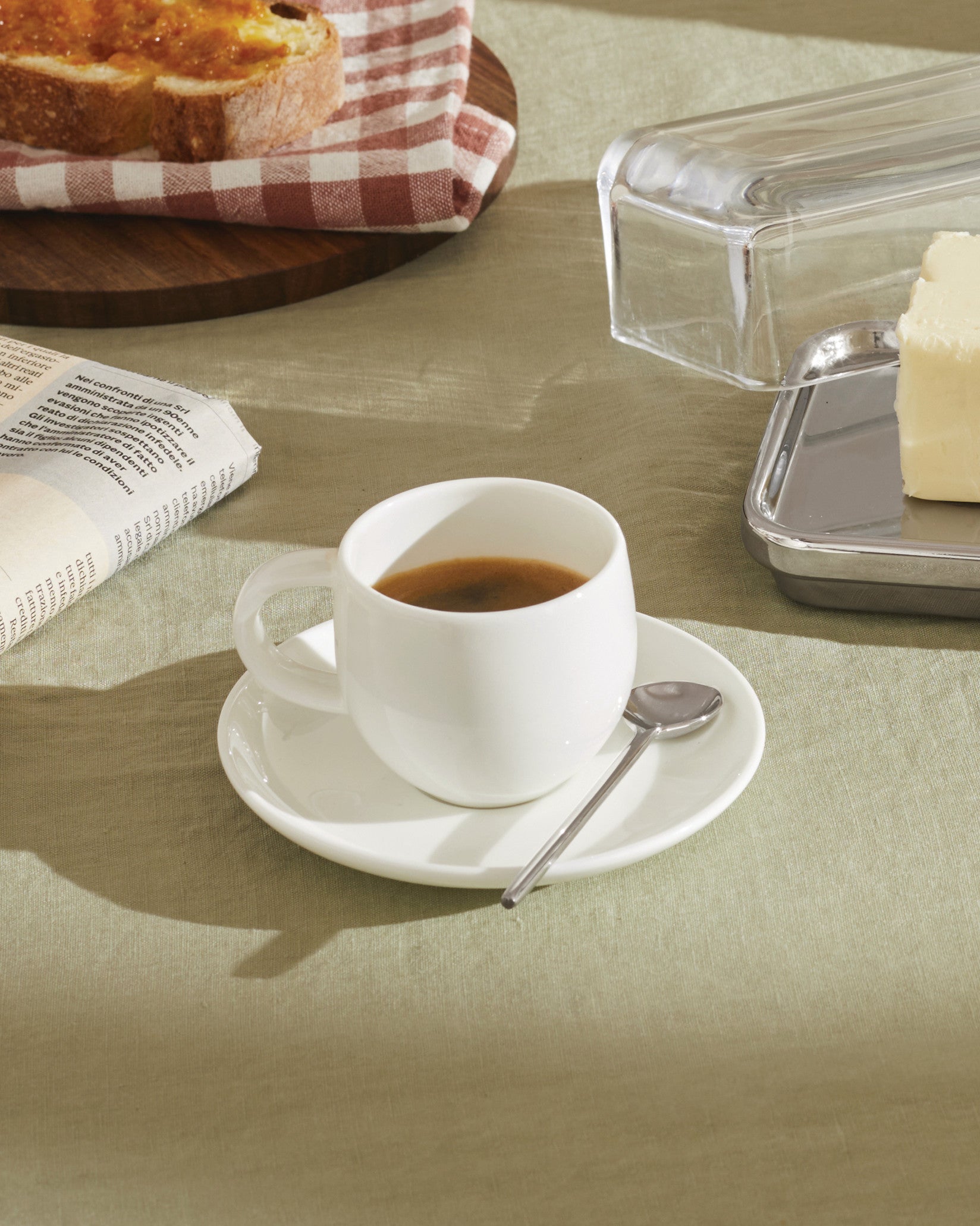 White ceramic espresso cup and saucer set by Alessi, with a spoon, on a light green tablecloth.
