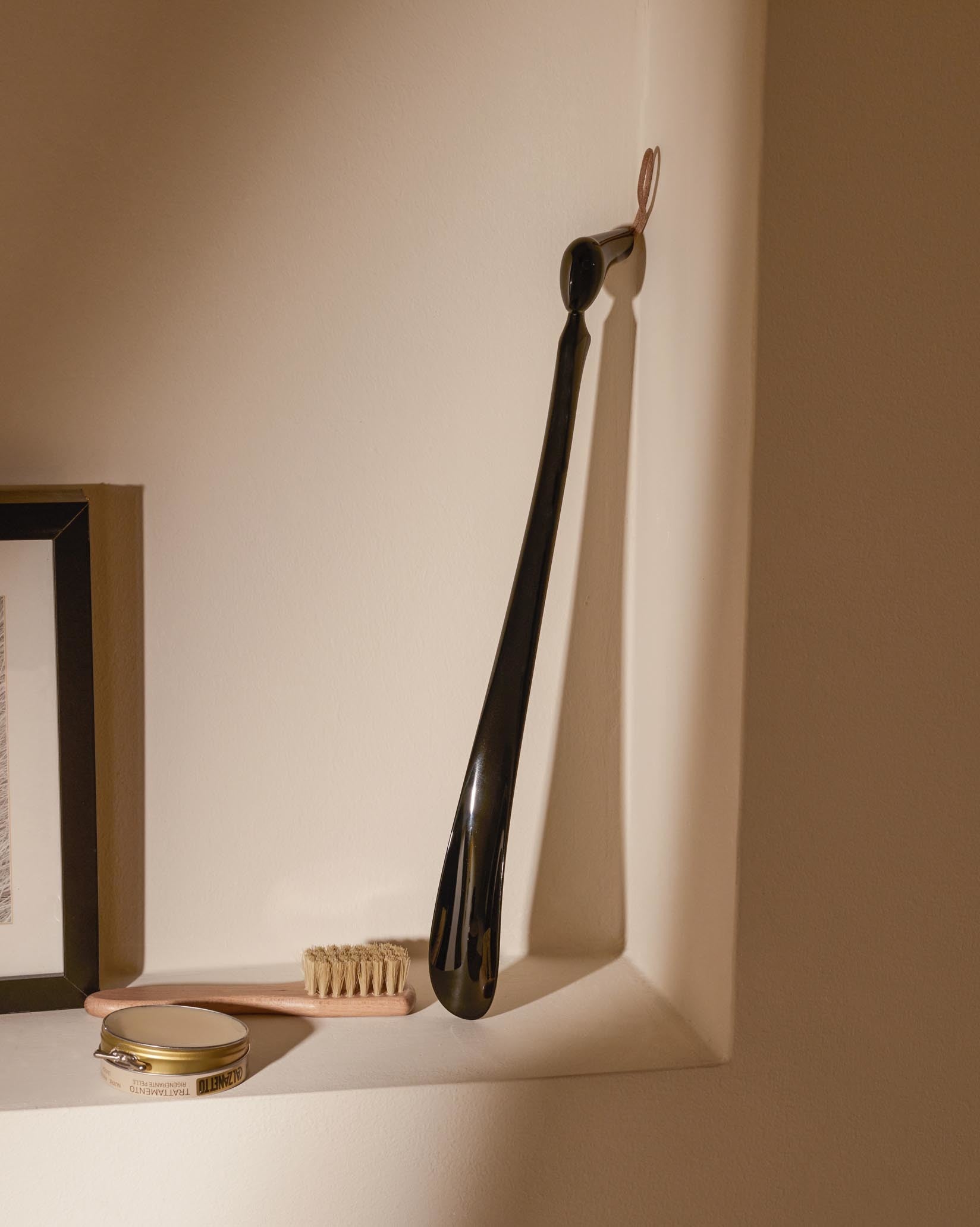 Black glossy shoehorn by Alessi with a brown leather loop, hanging on a wall above shoe polish and a brush.