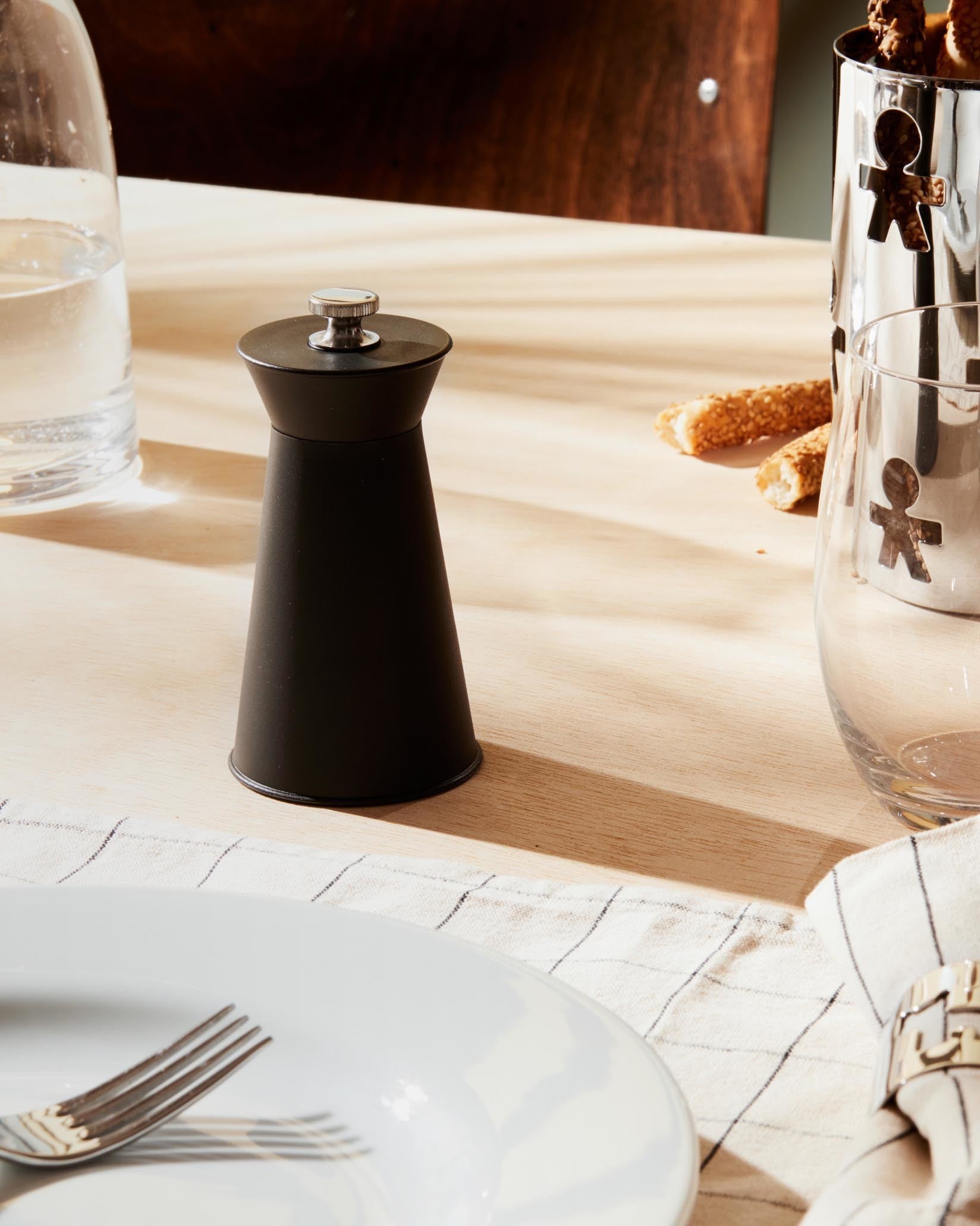 Matte black conical pepper mill by Alessi with a metallic knob, on a light wood table.