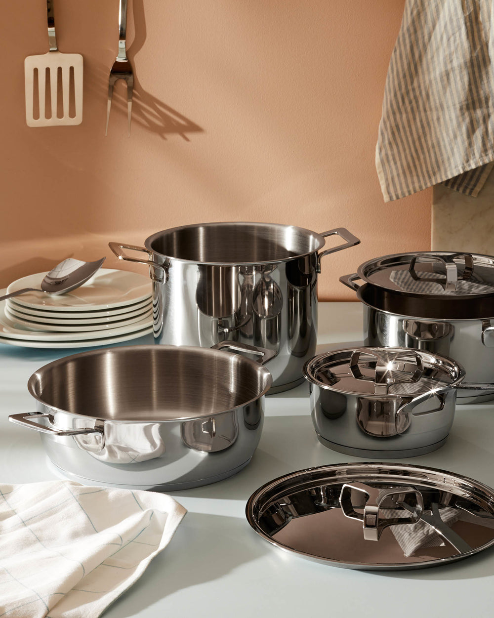 Shiny stainless steel pots and pans by Alessi, in various sizes with sleek handles and lids, on a light blue countertop.