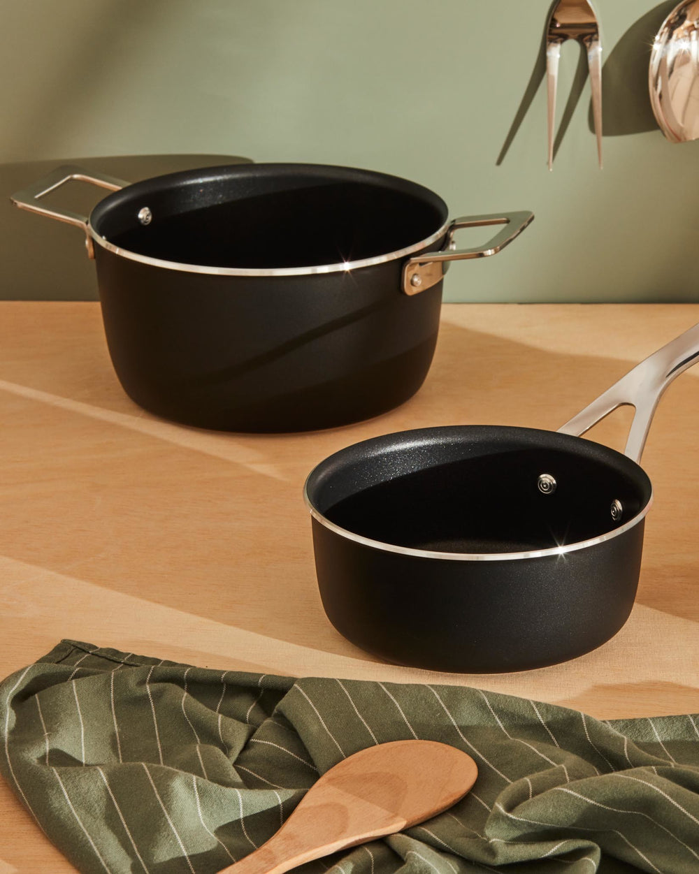 Black casserole pot and saucepan by Alessi with shiny silver handles and rims, resting on a wooden surface with a striped green towel.